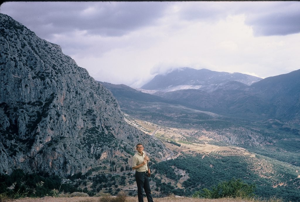 Scan of a Kodachrome slide, landscape, 1969. A young man in the near foreground in front of a long valley in Greece, with broken sun and weather clouds sweeping across it