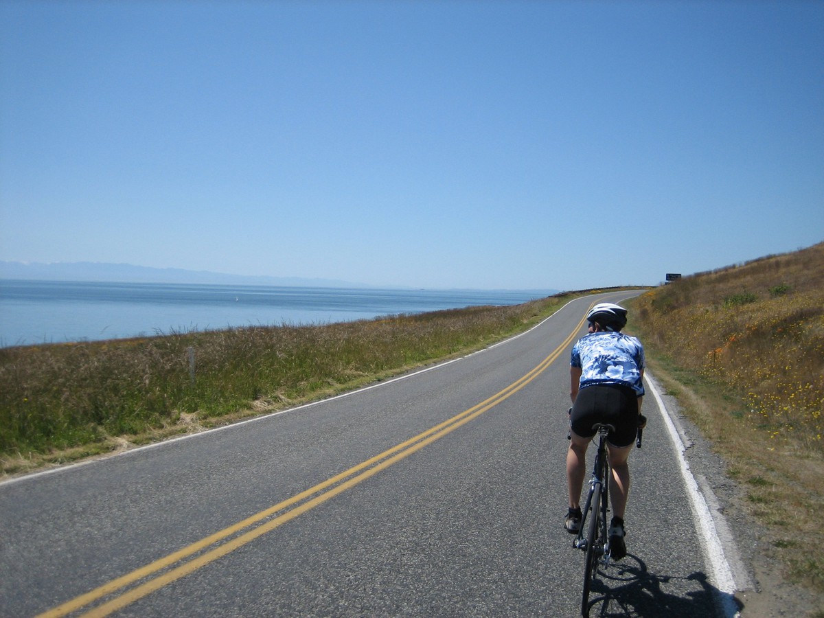 Outdoors, taken from a moving bicycle: a narrow empty highway along the seashore; a young woman on a road bicycle just ahead and moving our direction. Brilliant clear sunny weather