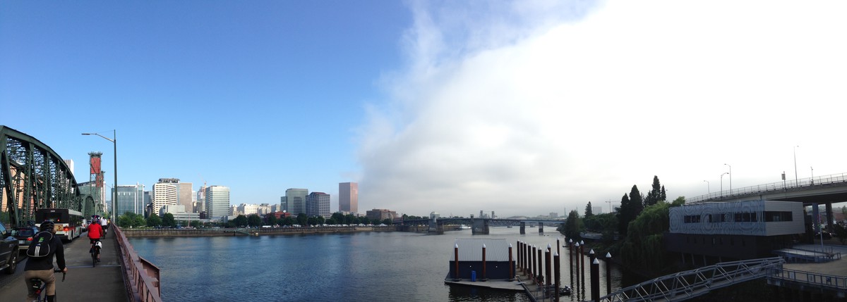 View up the Willamette River toward downtown Portland on a summer morning. To the left/west the sky is clear and sunny; to the right/east the sky is cloudy and foggy. This weather is neatly divided by the river itself