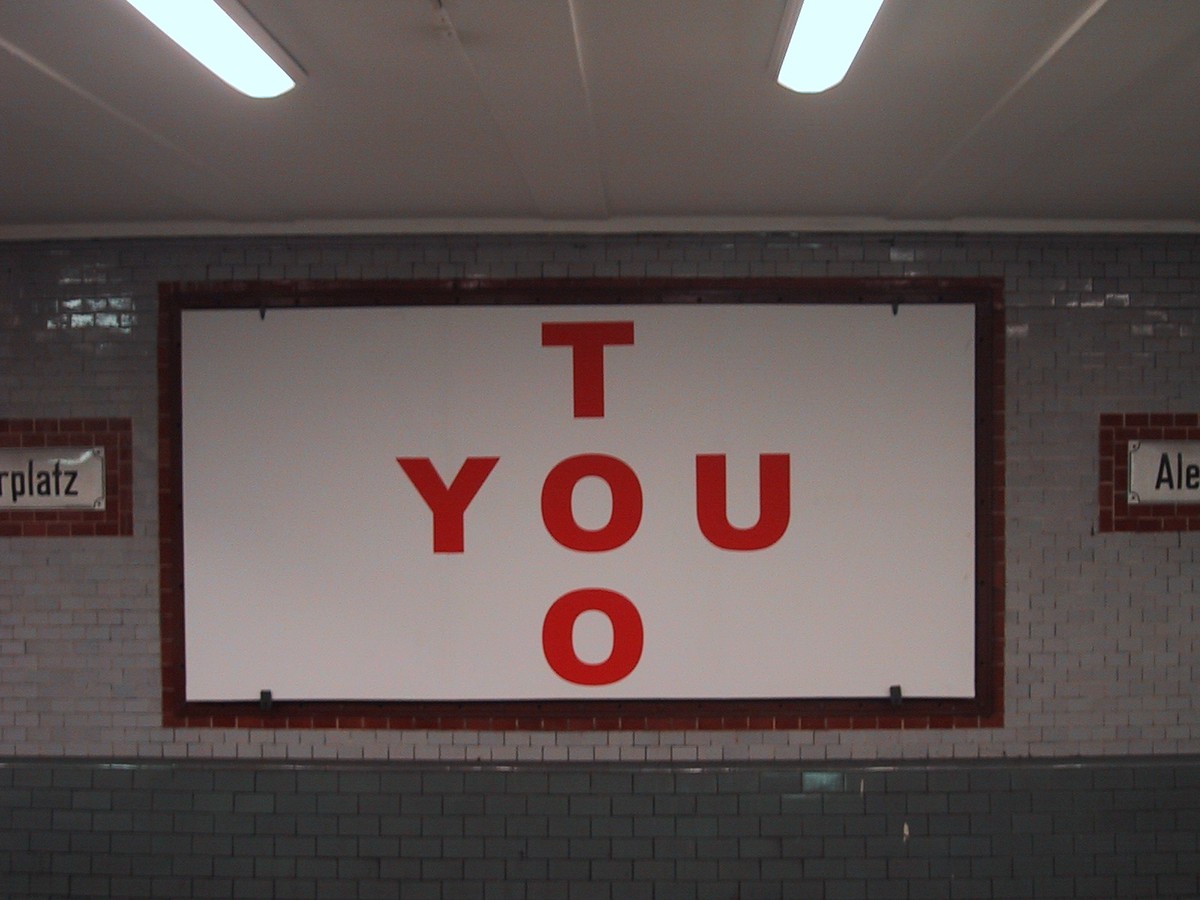 Simple billboard in a Berlin u-bahn station, reading “YOU TOO” in large block letters such that the Os in both words intersect, forming a cross shape