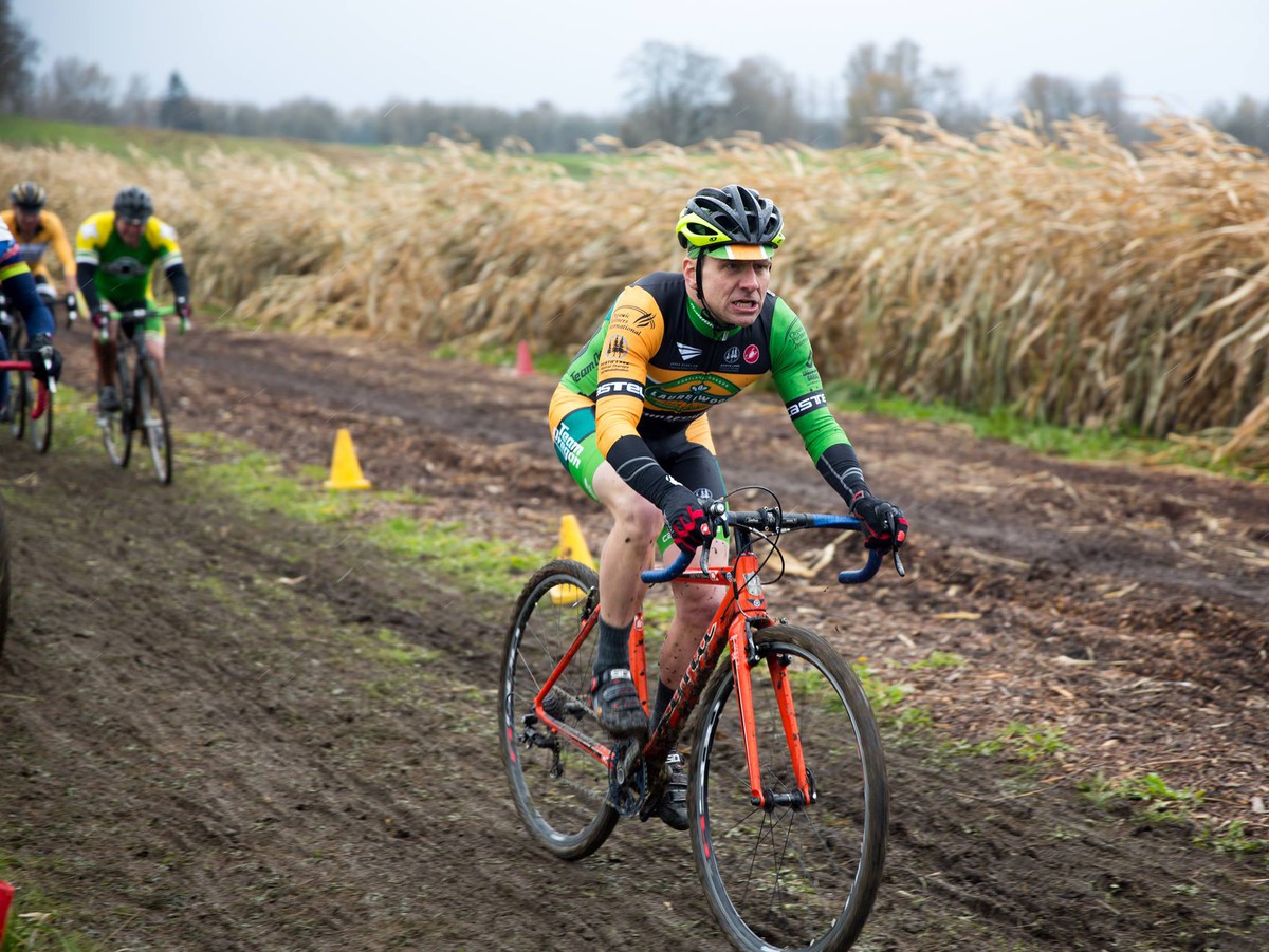 Middle-aged man on a cyclocross racing bicycle, wearing a colorful racing skinsuit, riding down a muddy track next to a field