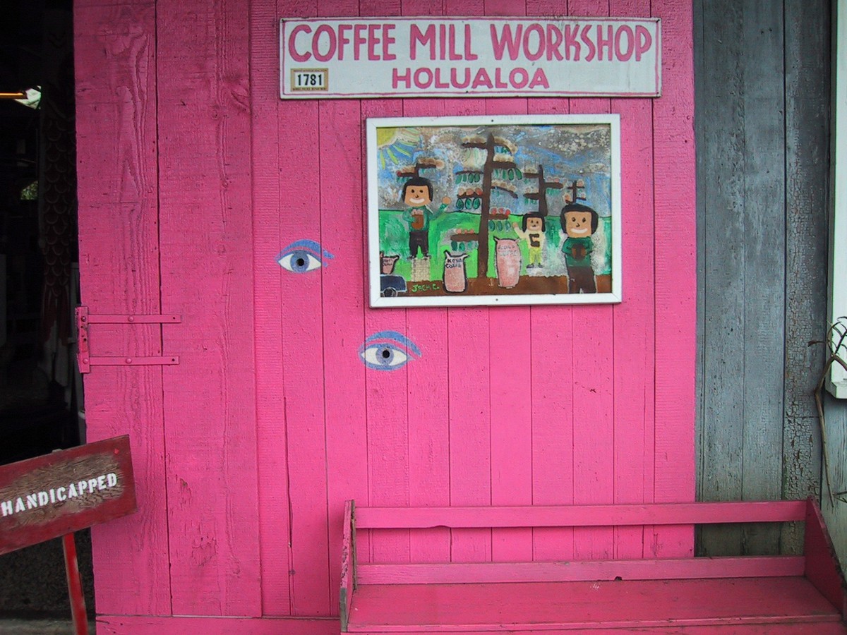 Empty bench in front of a colorfully-painted wooden barn, sign reads “Coffee mill workshop, Holualoa”