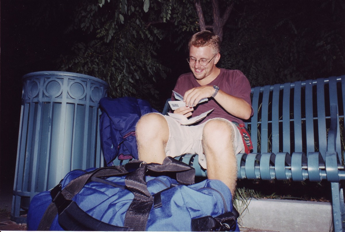 23-year-old white man, slightly drunk seated on a bench with a large duffel bag at his feet. Taken late at night, with a flash. He is looking at a train ticket