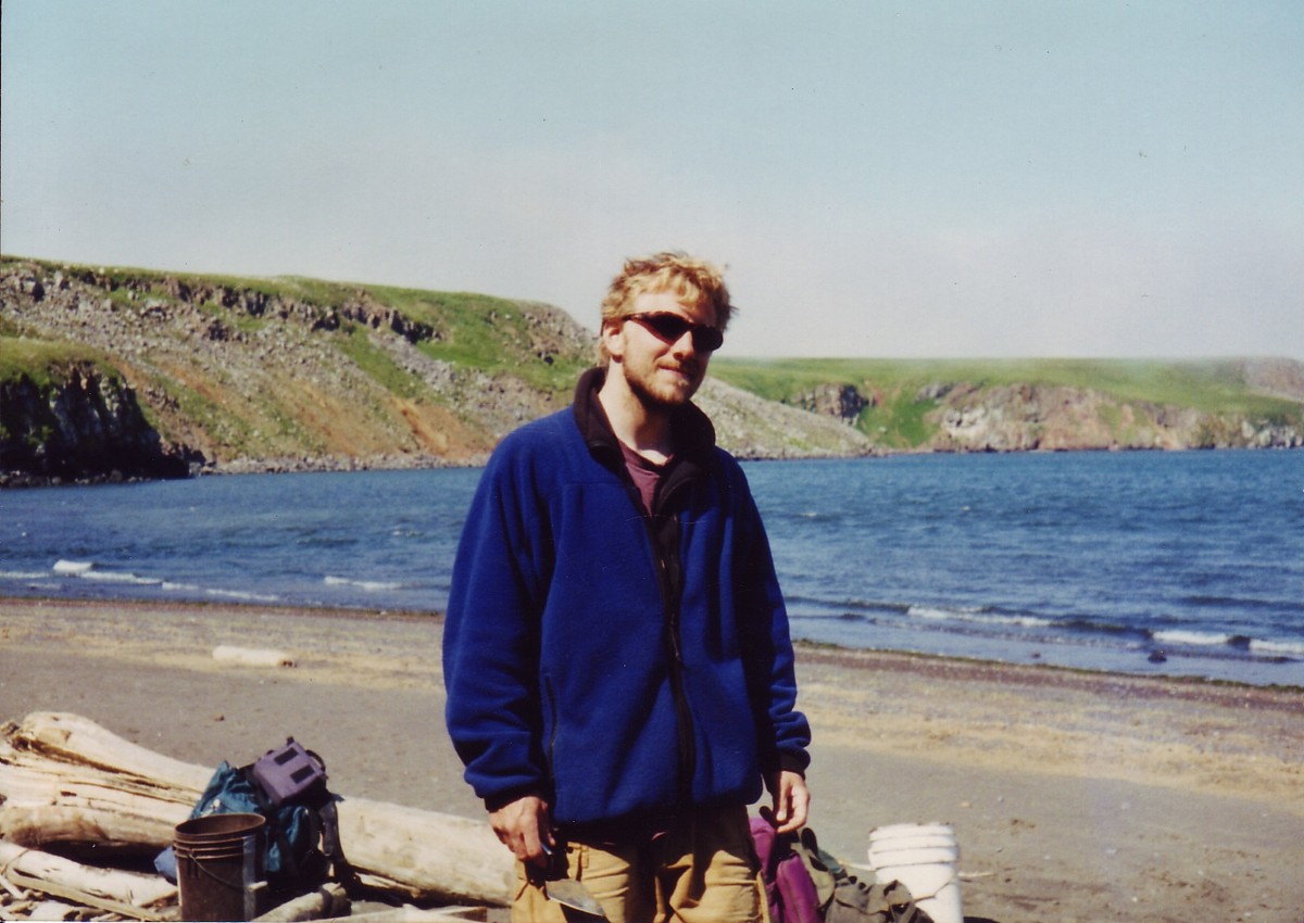 Casual portrait of a disheveled young man with a beard and sunglasses, wearing a dingy fleece jacket, holding a mason’s trowel. Taken on a sunny day on beach in the subarctic, with green cliffs across a narrow bay in the background