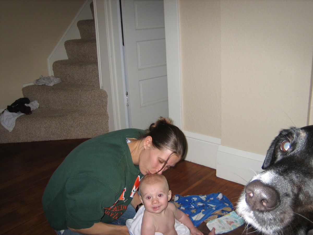 Indoors, a woman seated on the floor, kissing a baby on his head. Baby is wrapped in a towel. In the foreground a dog’s snout is visible just in the edge of the frame