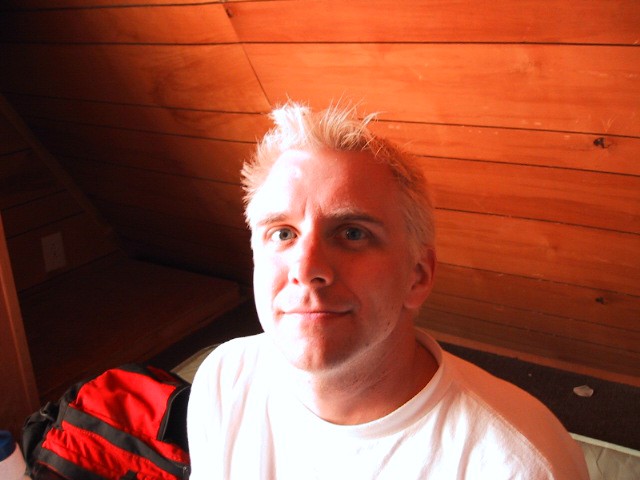 Moderate closeup, indoors in a rustic cabin, a young man with short bleached white hair making a cryptic smile