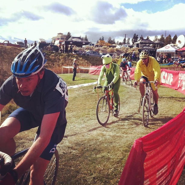 Action shot from a cyclocross bicycle race. In the center are two racers in costume, one is wearing a yellow jacket of some kind and the other is dressed as Kermit the Frog