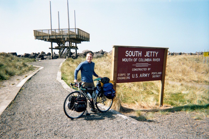 Casual portrait, outdoors, of a young man with a touring bicycle next to a sign that reads “South Jetty — Mouth of the Columbia River — constructed by U.S. Army Corps of Engineers Portland District”
