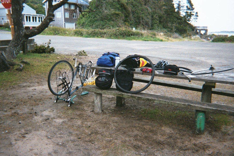 Picnic table next to a small parking lot at a coastal wayside area. A bicycle is upsidedown on its handlebars next to the table. All the touring gear is spread across the table; one wheel is off the bike, and the tire is off the wheel.