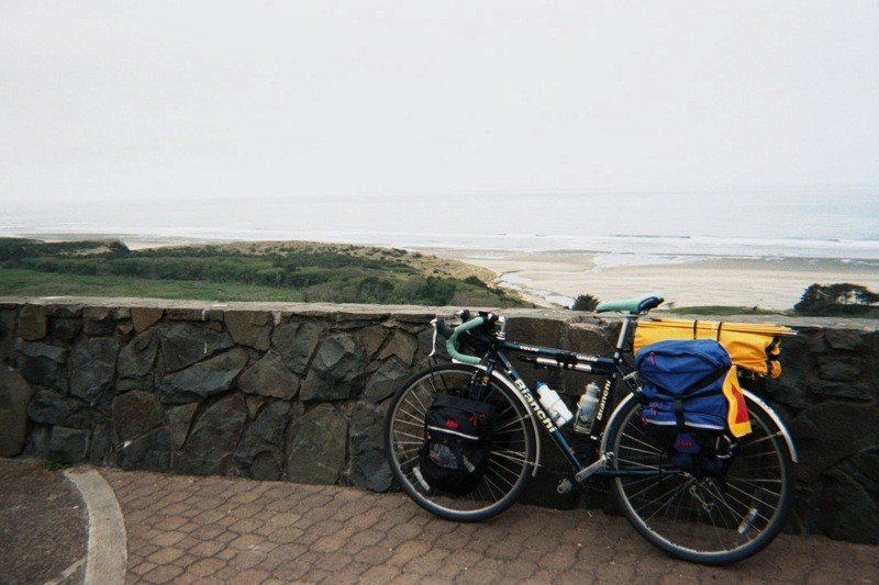 Loaded touring bicycle leaning against a low stone wall at a wayside viewpoint above a broad beach on the Pacific. Air is gray and damp.