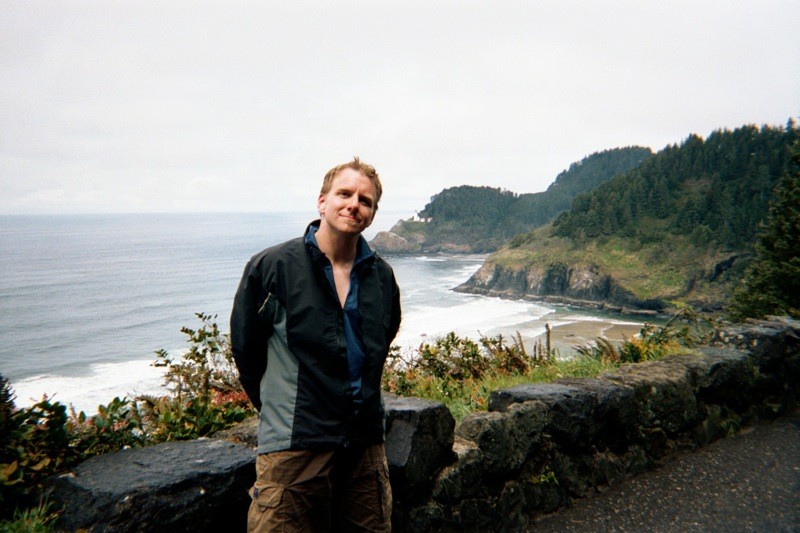 Landscape and casual portrait of a 30-year old man in wet bike gear standing at a roadside pullout on a seaside cliff with the ocean and two headlands in the background. The farthest headland is Heceta Head, with a lighthouse