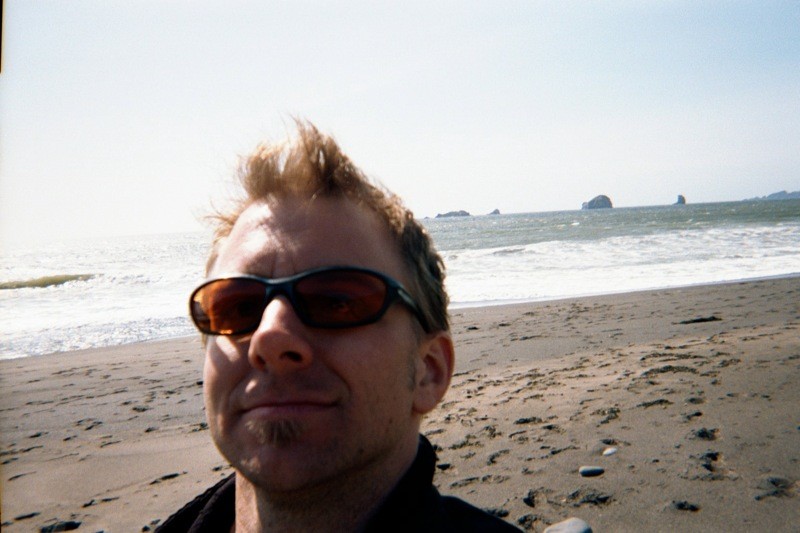 Selfie, white man with bikey sunglasses, on a windy beach facing west. Sea stacks a few miles offshore.