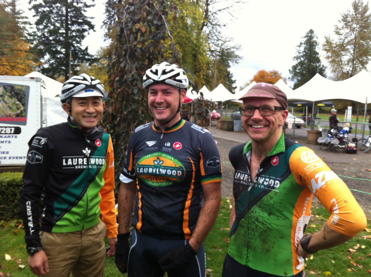 Casual portrait, three men in their late 30s/early 40s wearing coordinated “Team Oregon presented by Laurelwood” bicycle racing kit, next to a wet bicycle racing venue