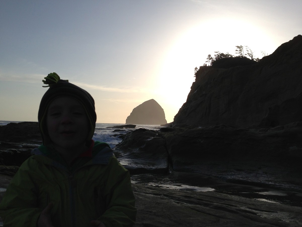 Young child facing the camera on a stormy (but clear) Oregon beach in winter. The sunset is blocked by a headland and sea stack; objects in the foreground are barely discernible as dark lumpy objects