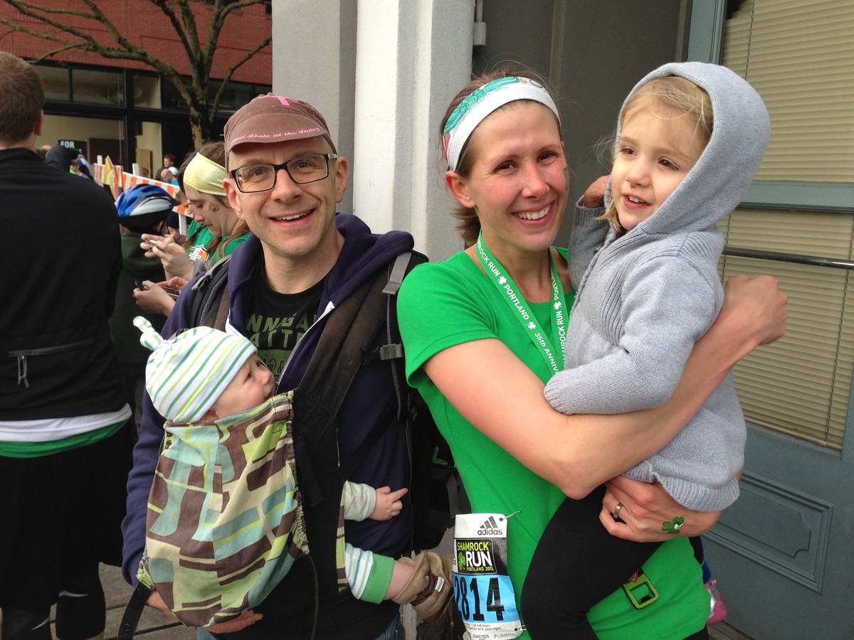 Casual portrait of a young family taken just after the finish at a marathon. The man wears a baby carrier with a young infant. The woman is wearing green running gear and a marathon bib; she holds a preschool age child