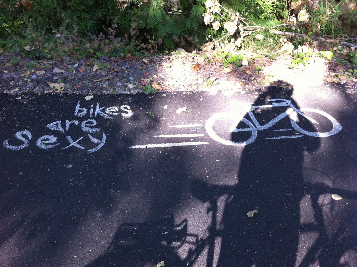 Graffiti on a bike path that reads “Bikes are Sexy” with a drawing of a bicycle. Taken with the sun behind so shadows of the photographer and bicycle are visible