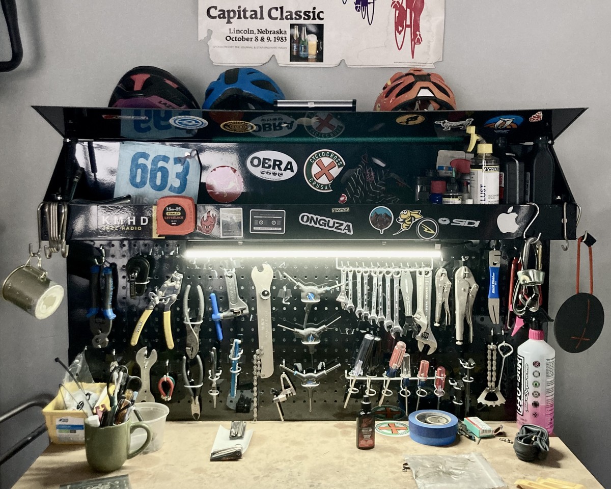 Workbench in a home bike shope with magnetic pegboard, thick with a motley assortment of tools. The workbench is covered with bicycle and outdoor sports-related stickers