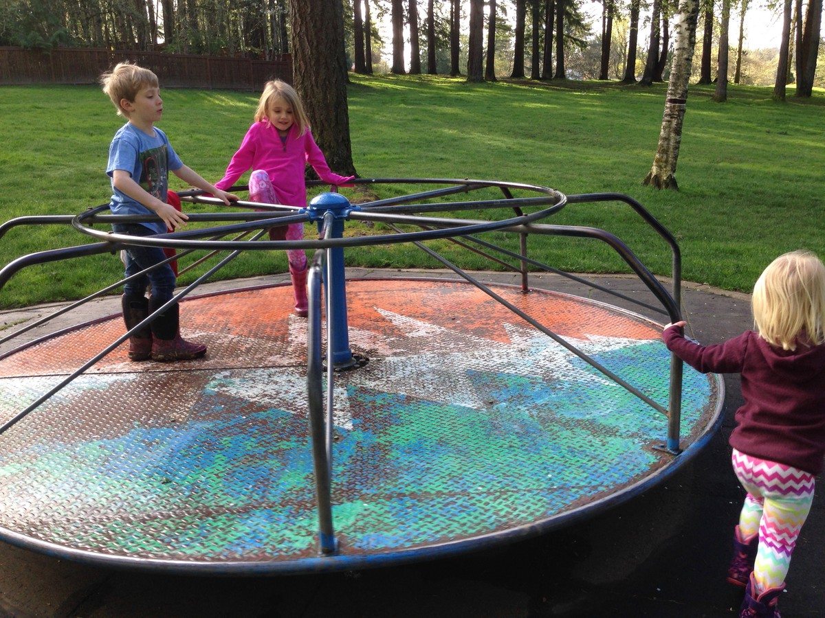 Three young kids with mudy boots, on a rusty old merry go round at a tree filled park
