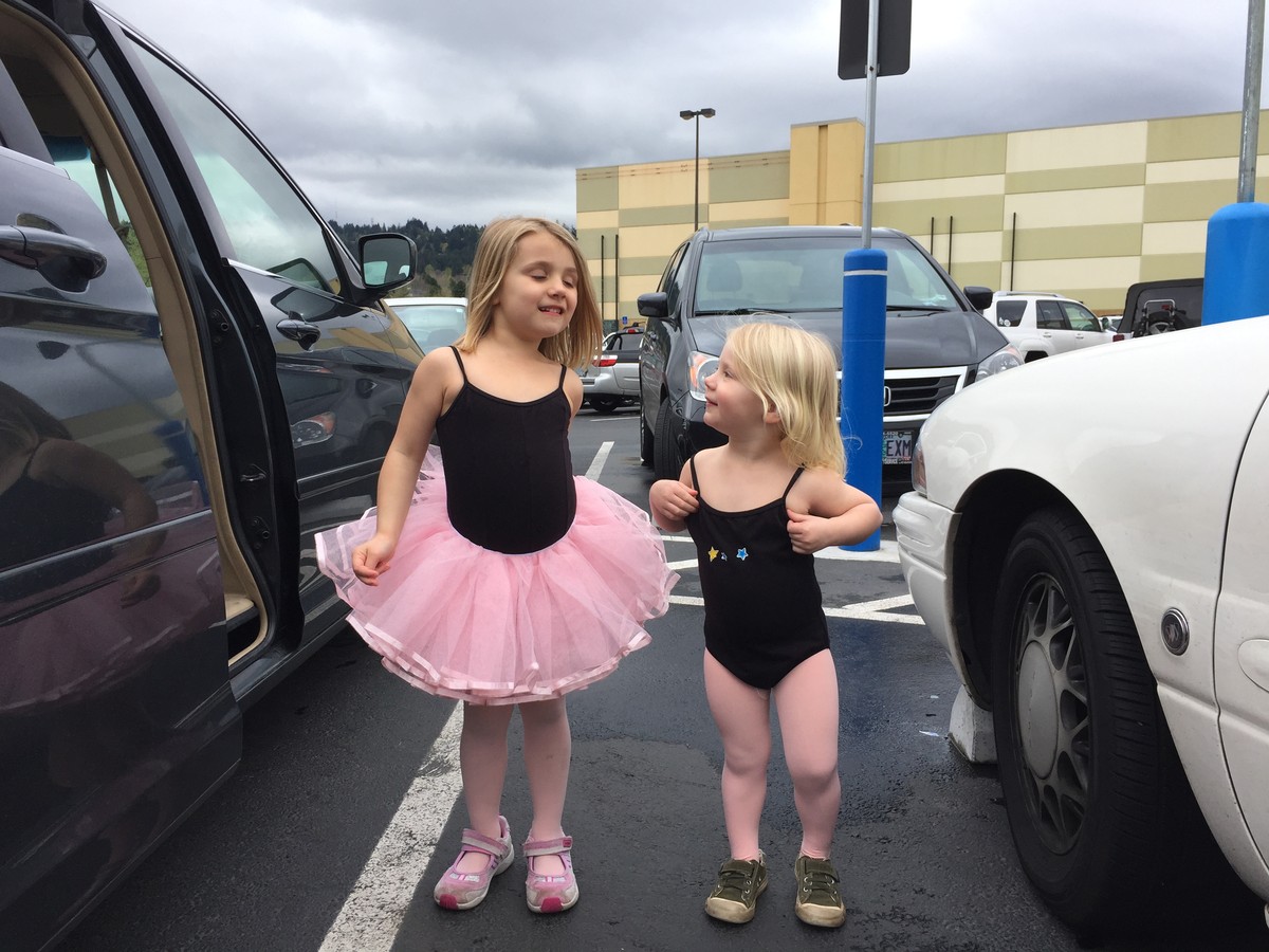 Two girls ages 5 and 3 in ballet leotards, in a parking lot. The smaller, younger girl is looking up at the older girl with some admiration