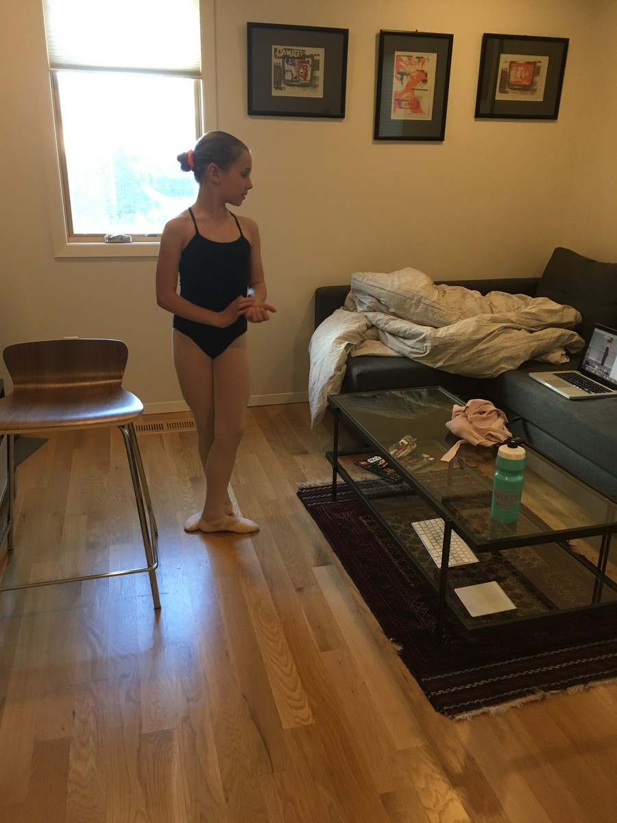 Indoors, TV den, school age girl in a leotard standing at first position, watching an instructor on a laptop