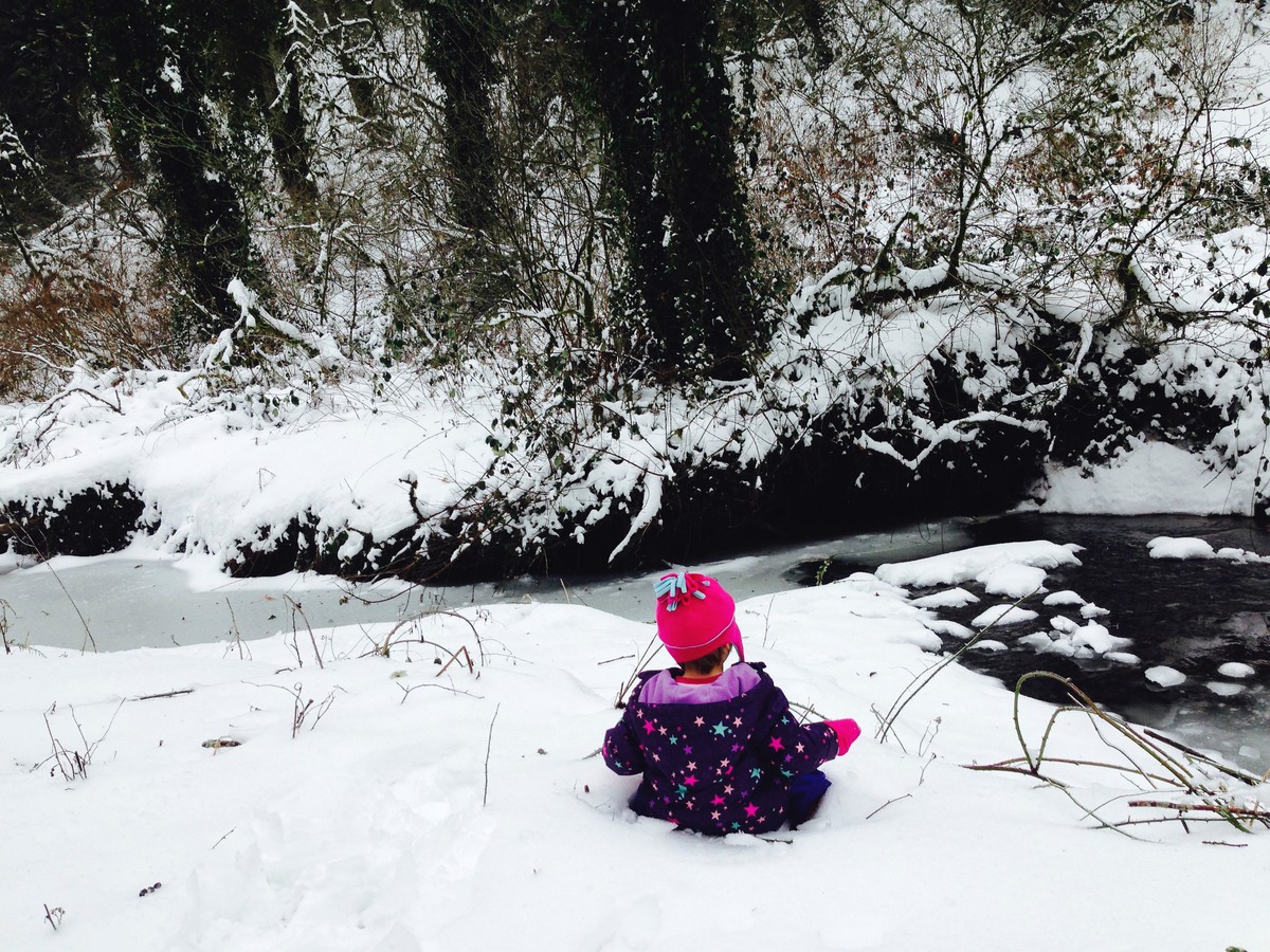 Preschool child in colorful snow gear sitting in the snow next to a partially-icy stream in the forest