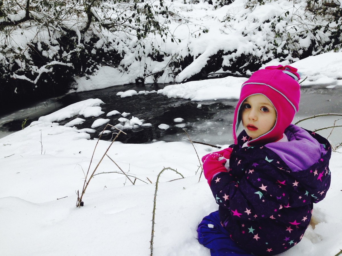 Preschooler in colorful snow clothes seated by a (flowing) river in a snowy forest; she is looking over her shoulder toward the camera