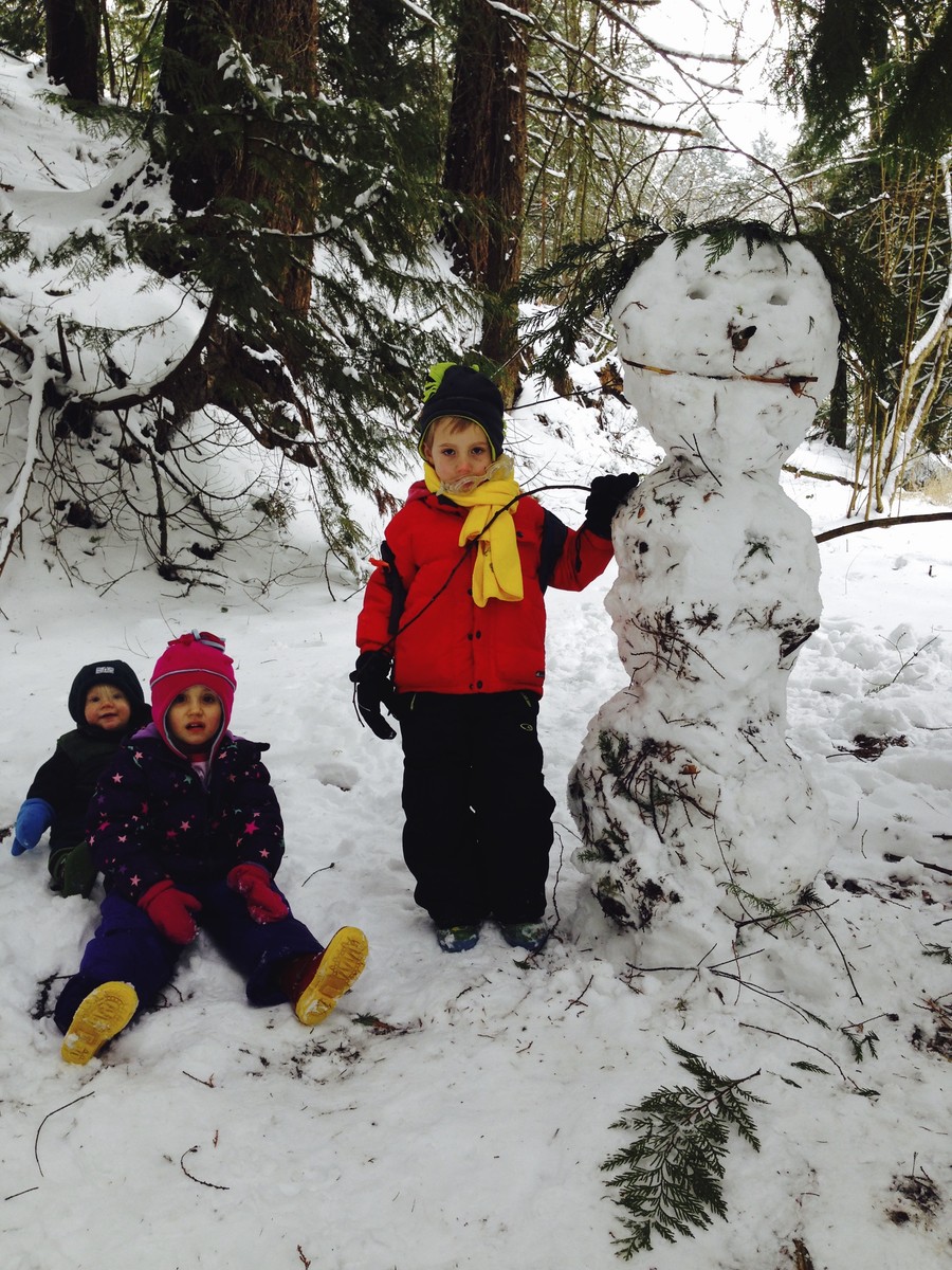 Three small children in snow clothes next to a snowwoman, in a snowy forest