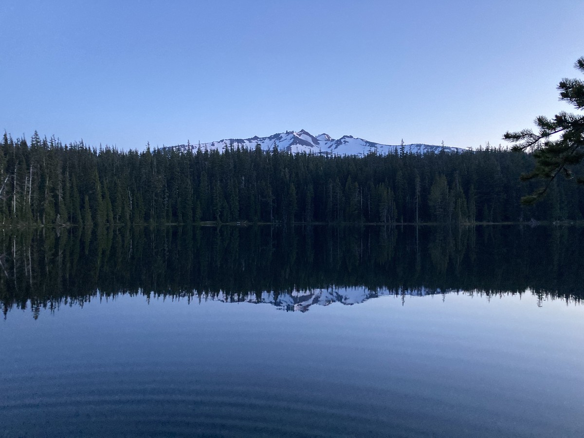 Landscape across a still forest lake at twilight. A single ring ripples slowly across the lake. On the horizon, looming above the trees on the far shore, is the glaciated peak of a broad razorback mountain. Excepting the ripples the lake presents an almost perfect mirror of the scene