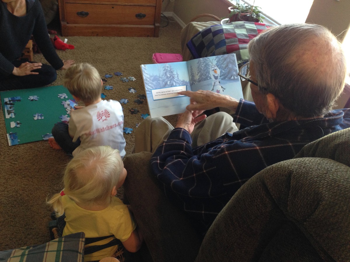 View over the shoulder of an old man reading a FROZEN picture book to a very young toddler. A woman and a young school age boy are putting together a FROZEN puzzle on the floor
