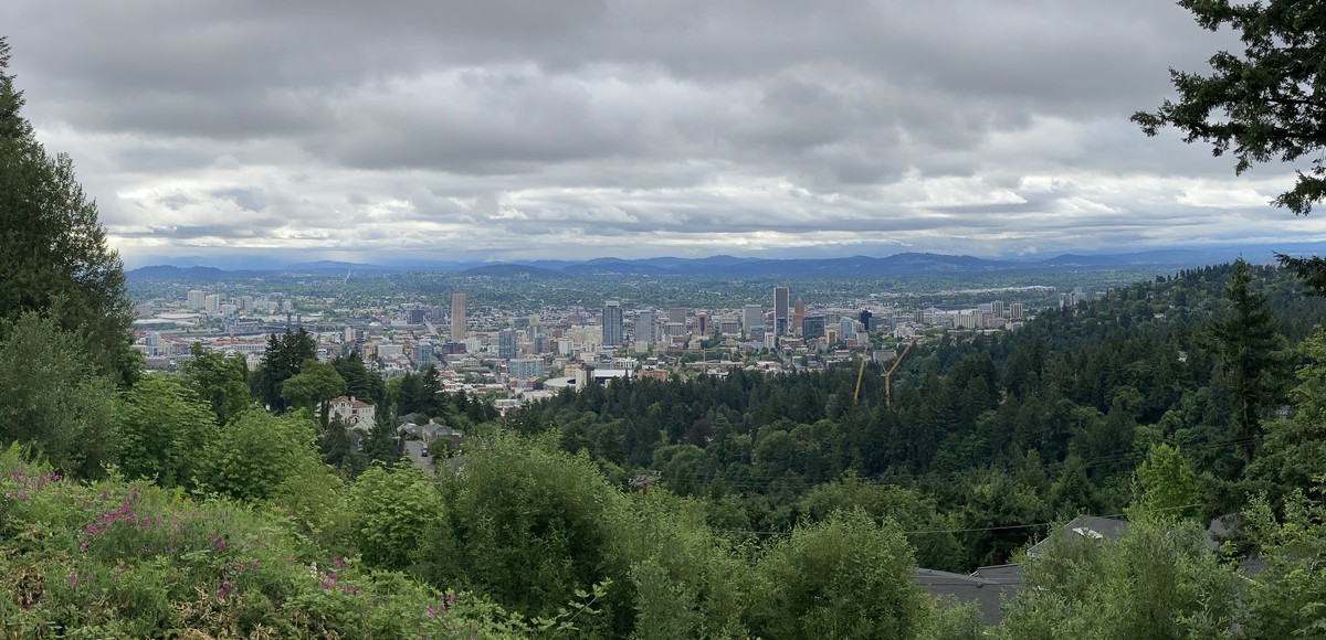 Panorama of downtown and inner eastside portland, taken from Pittock Mansion looking east. A high deck of clouds obscures the far horizon but the air is clear and visibility good. The city is lush with early summer foliage and looks clean and inviting