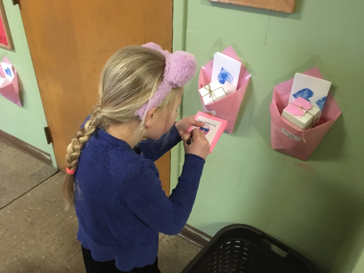 Kindergartener with braids and fuzzy animal ear headband hastily scribbling on Valentine’s day cards outside a classroom