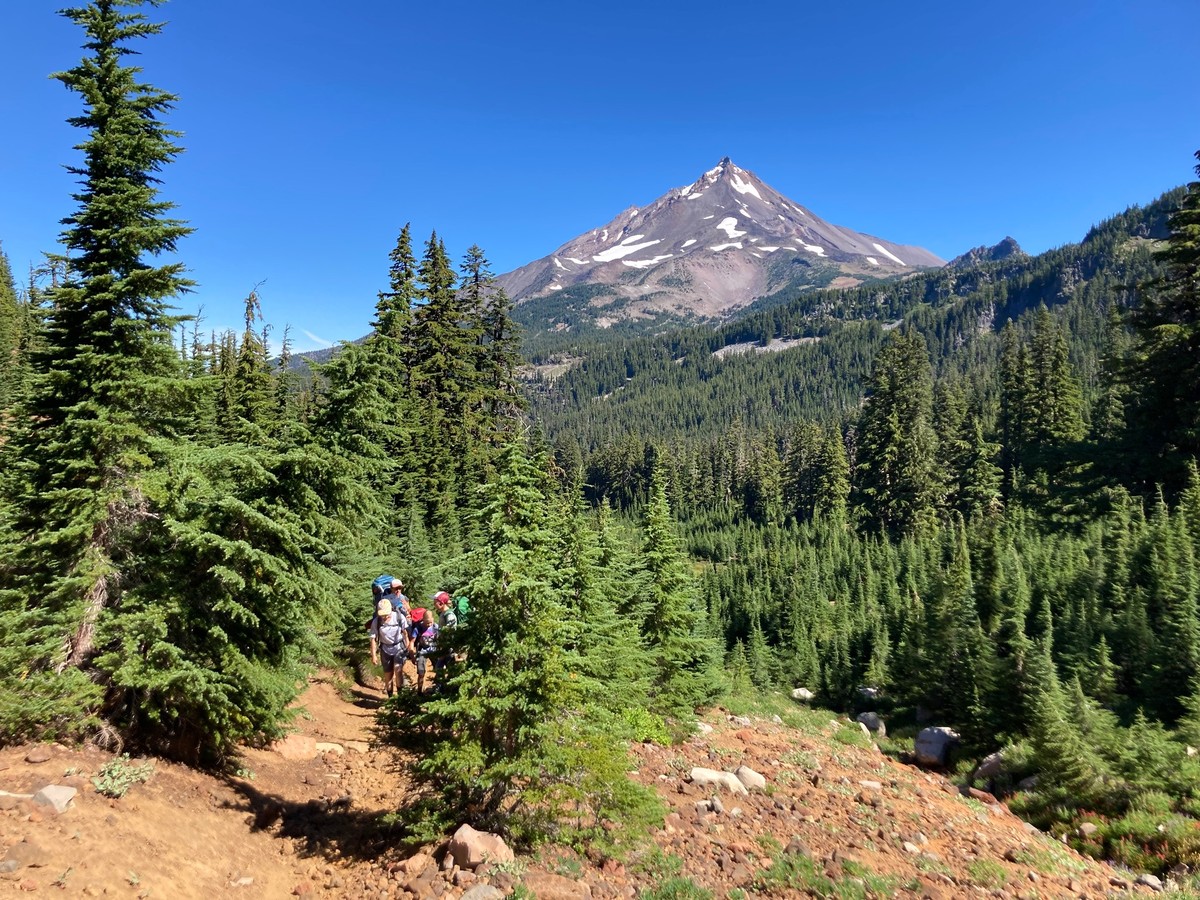 Four backpackers in shorts climbing a trail out of a broad alpine valley, with the triangular peak of Mt. Jefferson in the background, under a sparkling clear sky