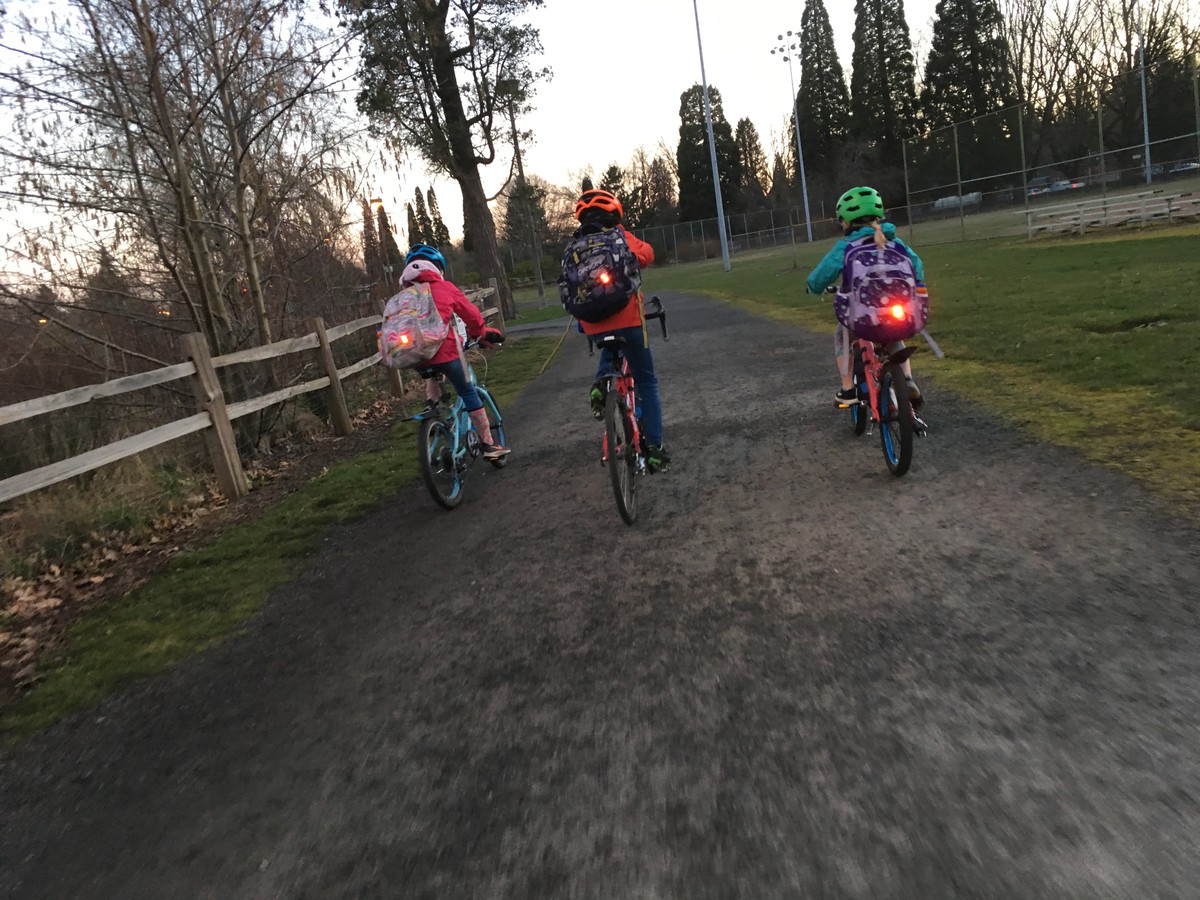 Back view of three school-age children on bicycles, with backpacks and blinky red tail lights, riding up a gravel path in a park