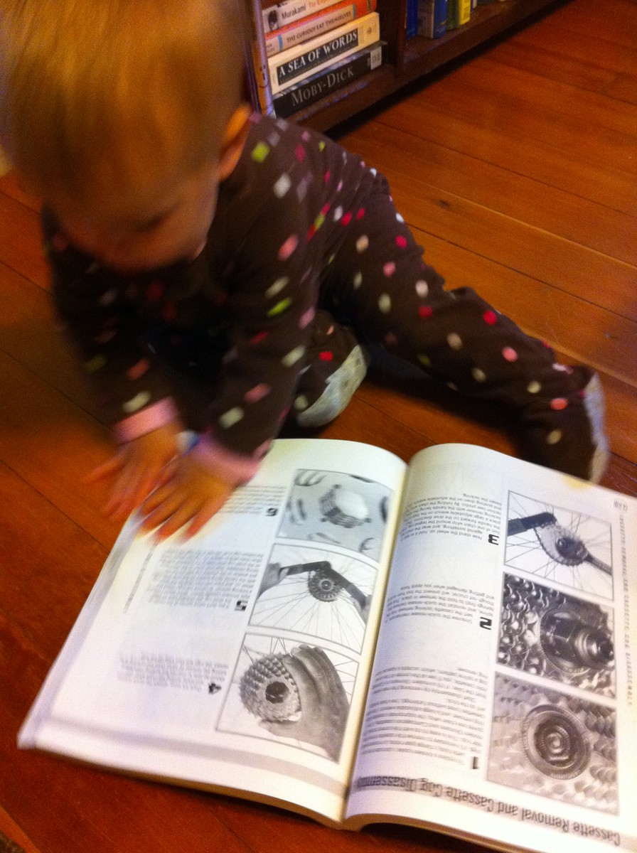 A baby in footie jams seated on the floor paging through an illustrated bicycle repair manual