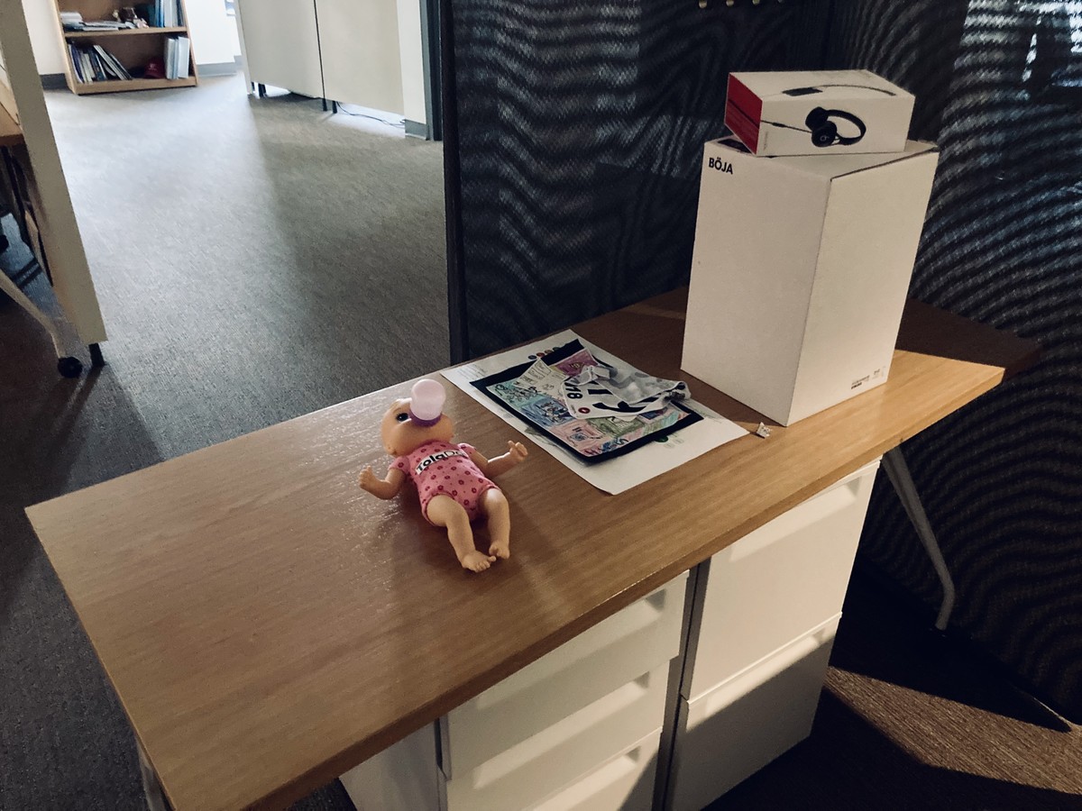 A mostly-bare desk in a darkened, empty office. 2 notable groups of objects on this desk: 1. a plastic baby doll marked “TolaData,” 2. a small pile of personal photographs & effects