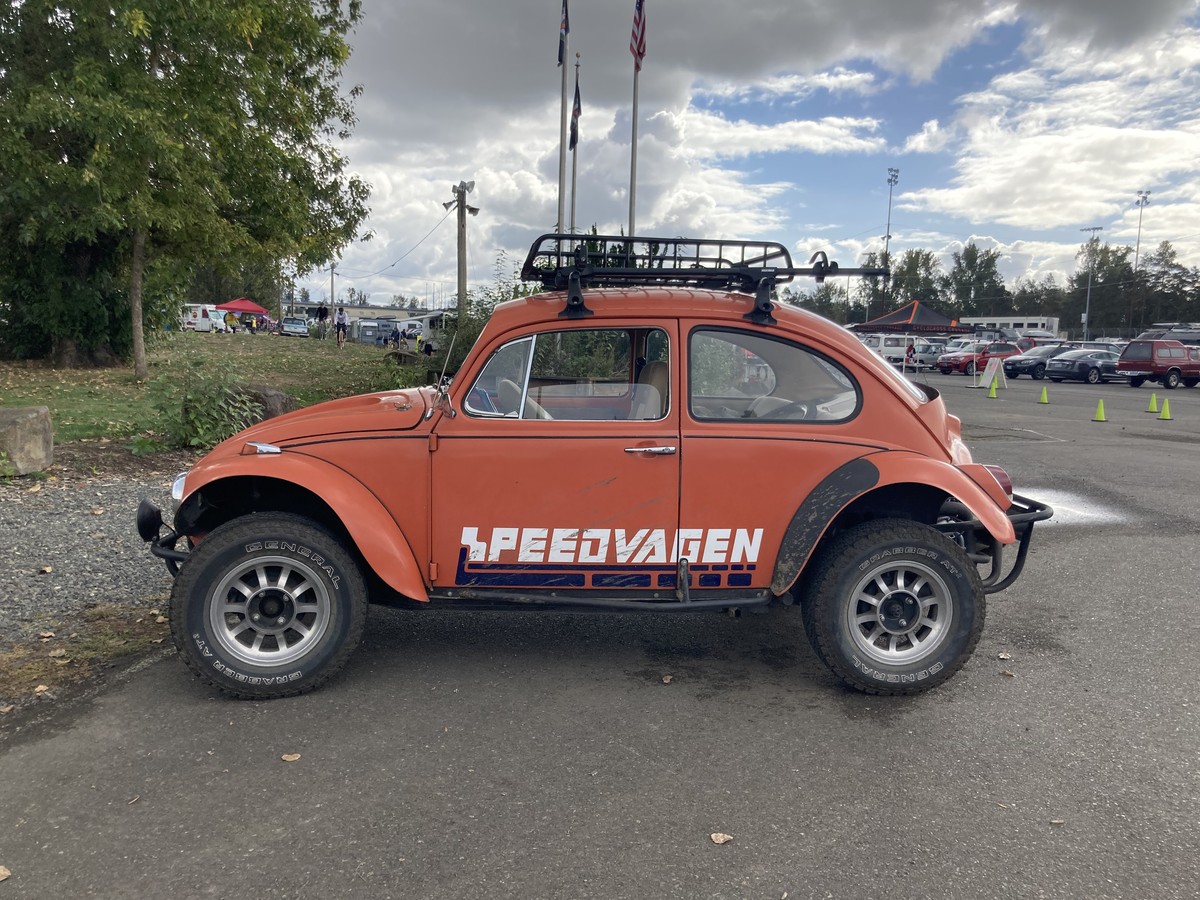Driver’s side a VW Beetle, Baja mod, with “Speedvagen” painted on the side, and a roof rack. Parked near to the registration tents at a bicycle race