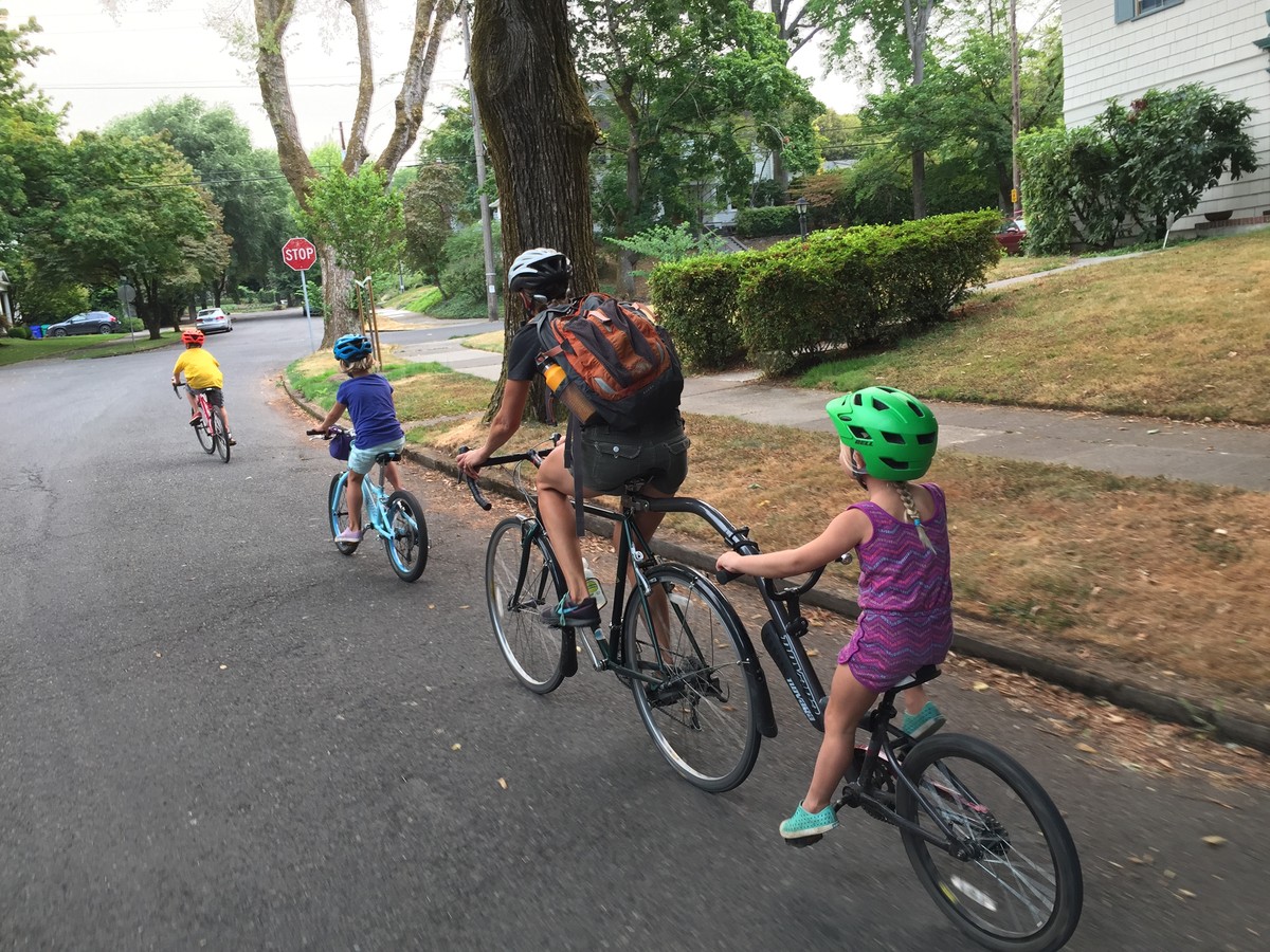 Two kids on kids’ bikes, a woman on a touring bike, towing a third kid on a tagalong bike