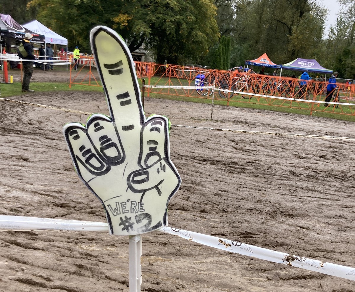 handmade sign at a very muddy cyclocross race, showing a raised middle finger and the text “we’re #2”