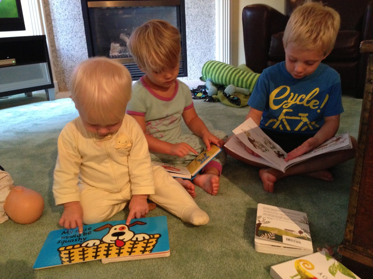 Three very young children (ages 1–5) sitting on a living room floor looking at books