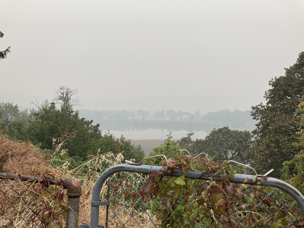Landscape taken across a rusty chainlink gate, from a bluff above a swampy lake. It is cloudless but visibility is less than 1 mile with a heavy smoke from wildfires