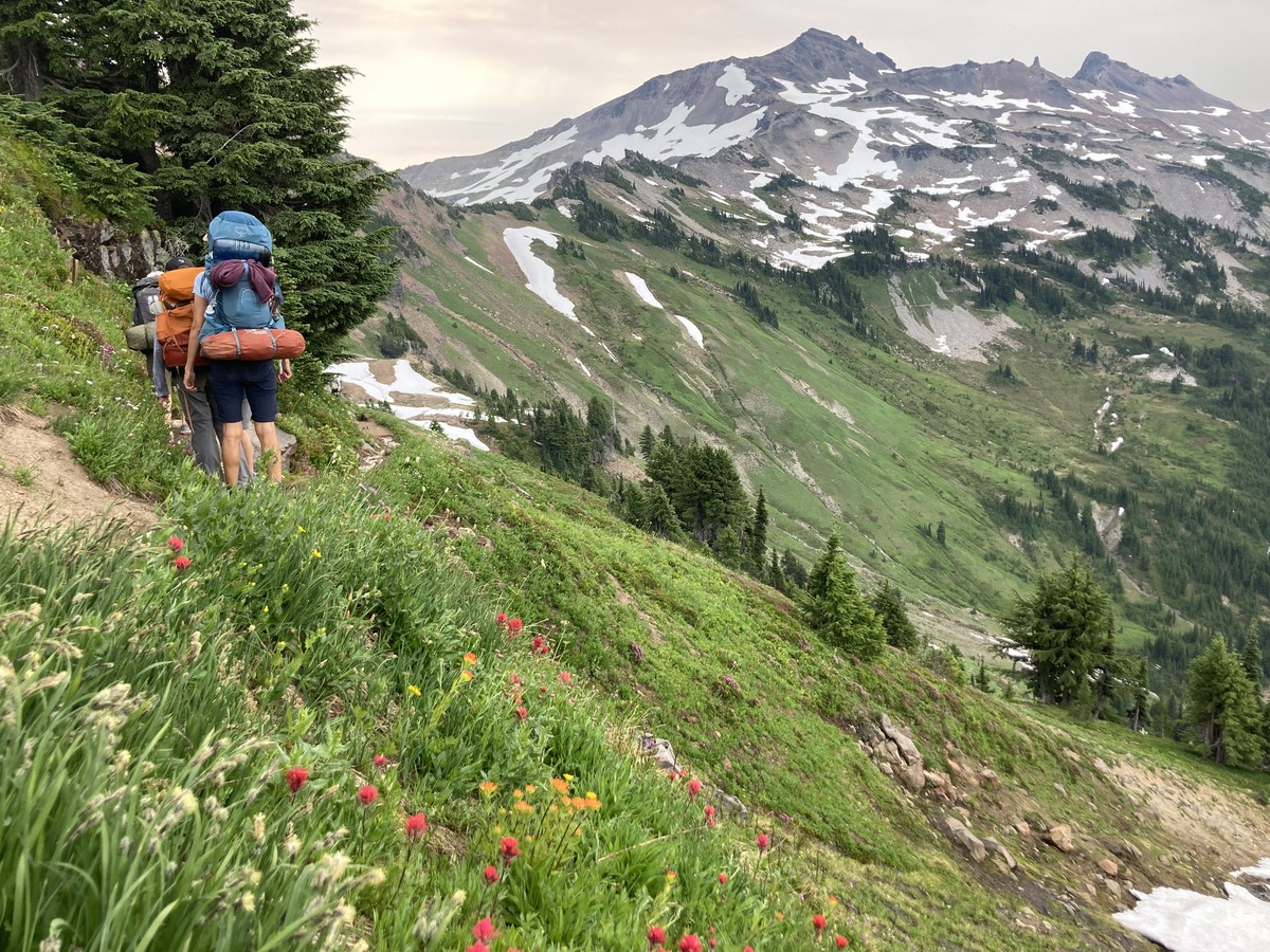 A group of backpackers climbing a narrow path up an alpine ridge toward a cluster of mountains dotted with patches of snow