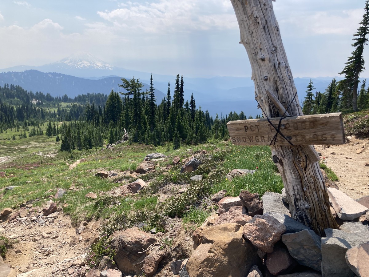 A heavily repaired trail junction sign and cairn in the foreground on a rocky alpine meadow with a stand of firs in the near distances. Mt. Adams on the horizon, its peak cloaked in haze and clouds