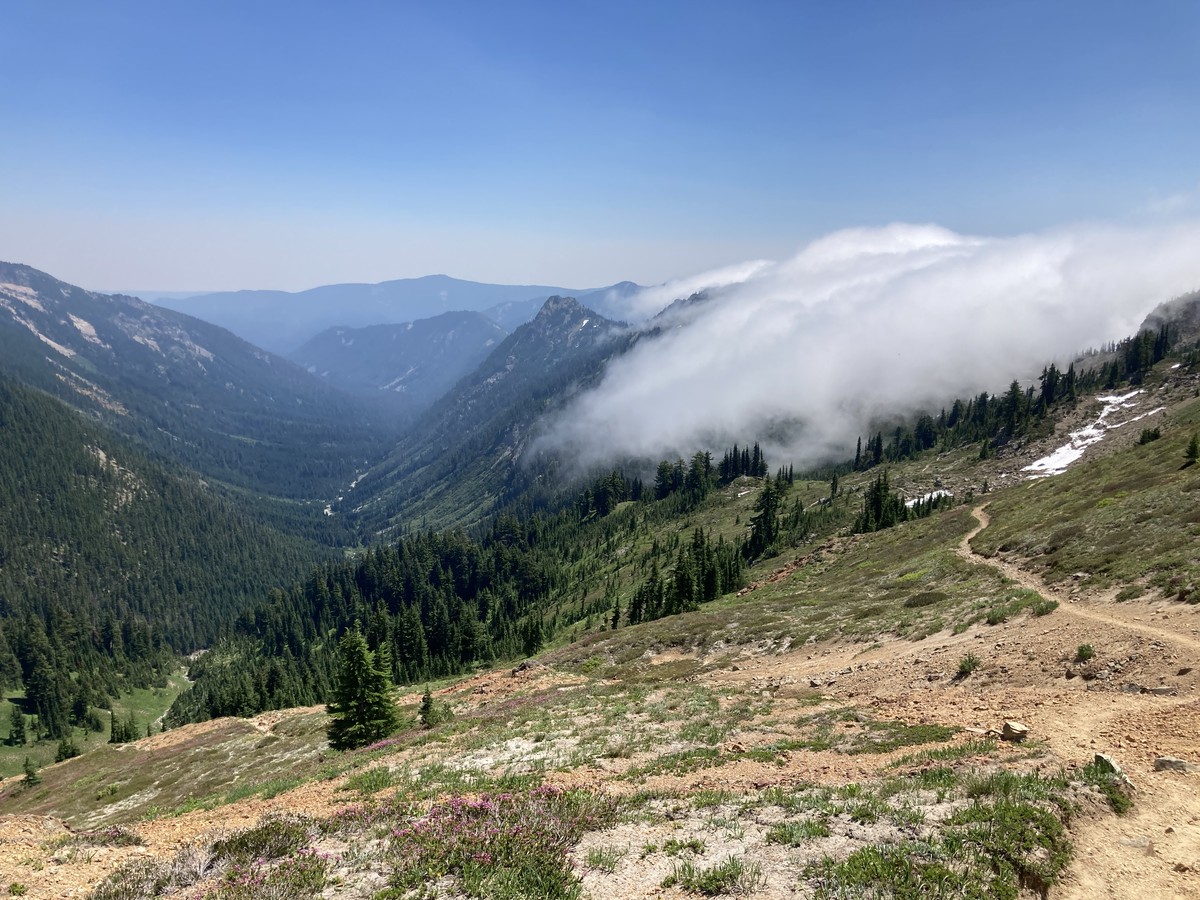View from the top of a barren alpine pass looking down a u-shaped glacial valley toward forested mountains. On the right/west, a thick fog of marine clouds rolls over the ridge from the neighboring valley