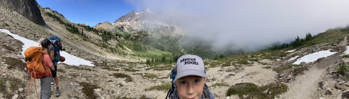 Panorama of a high alpine valley with a young person in the foreground, looking into the camera, wearing a STREET ROOTS cap. We are just at the boundary where a layer of marine clouds are rolling into the valley, experienced here as a heavy fog to one side and clear sunlight on the other