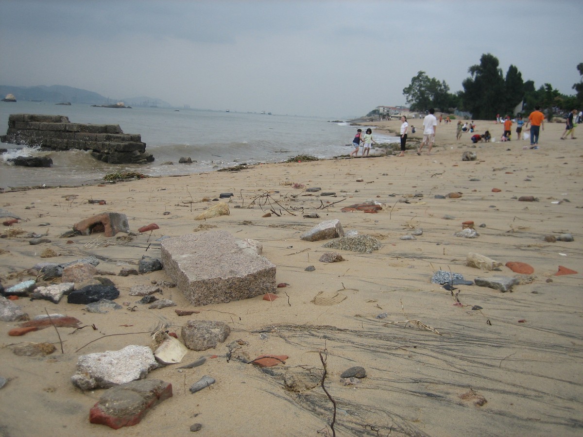 Low perspective shot up a beach with many highly-weather objects of architectural debris (bricks, glass, ceramic, rebar, concrete) in the foreground. About 200m distant are small groups of beachgoers and dogs