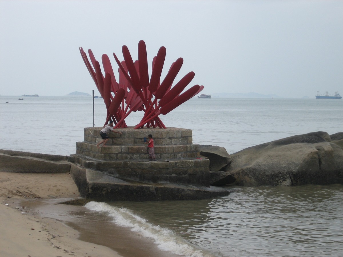 3-4m tall red metal sculpture resembling fans, on a stone and concrete plinth, with a person climbing up the plinth. The weather is gray and hazy; large cargo ships are visible on the ocean horizon behind