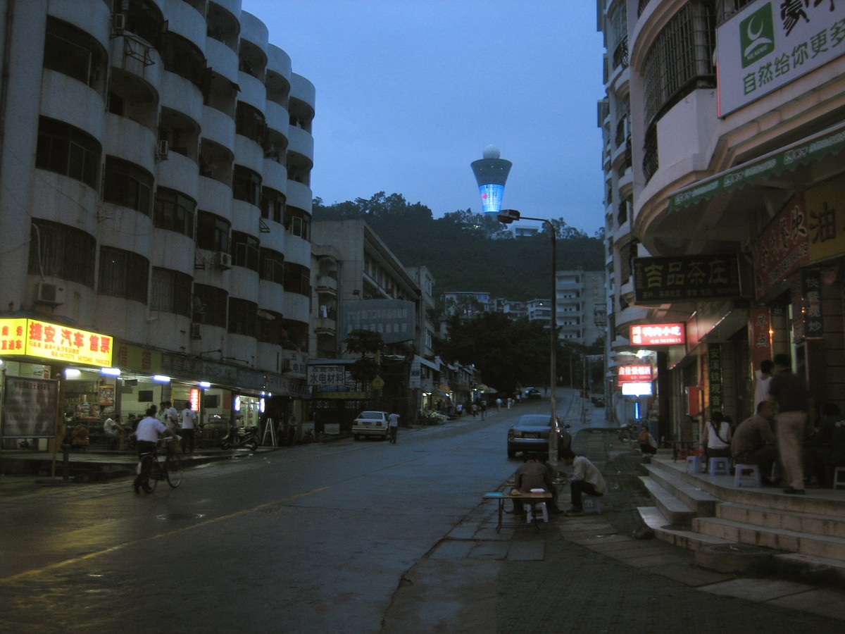View up a mostly-darkened street toward a brightly-lit neomodern tower on a forested hill. The buildings in the foreground are of an aging midcentury style once popular in China; they have active street life in the form of well-lit commercial shops, but the buildings are otherwise completely dark (and likely abandoned)