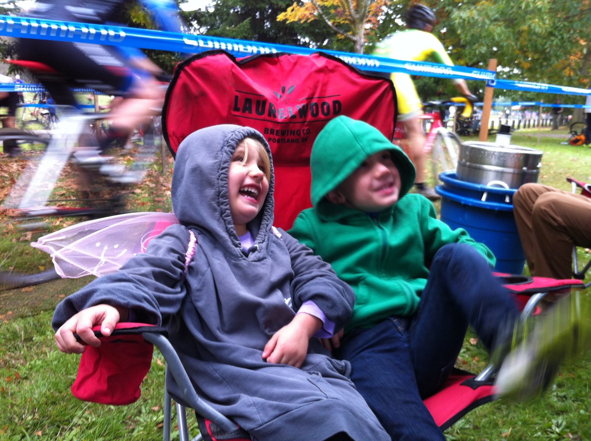 Two young children in sweatshirts (one wearing fairy wings) in a camp chair branded “Laurelwood Brewing Co.” at a bicycle race. They are smiling/laughing at someone off-camera. Very close behind them are two bicycle racers mid-race; to their collective left is a keg of beer in a huge ice bucket