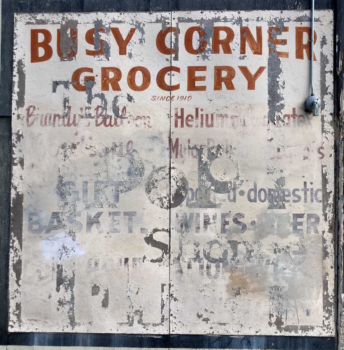 Faded commercial sign from a neighborhood convenience store, long-since closed, reading “BUSY CORNER GROCERY since 1910” followed by a mostly-illegible list of product that includes “Helium,” “Wine,” and “Gift Baskets”