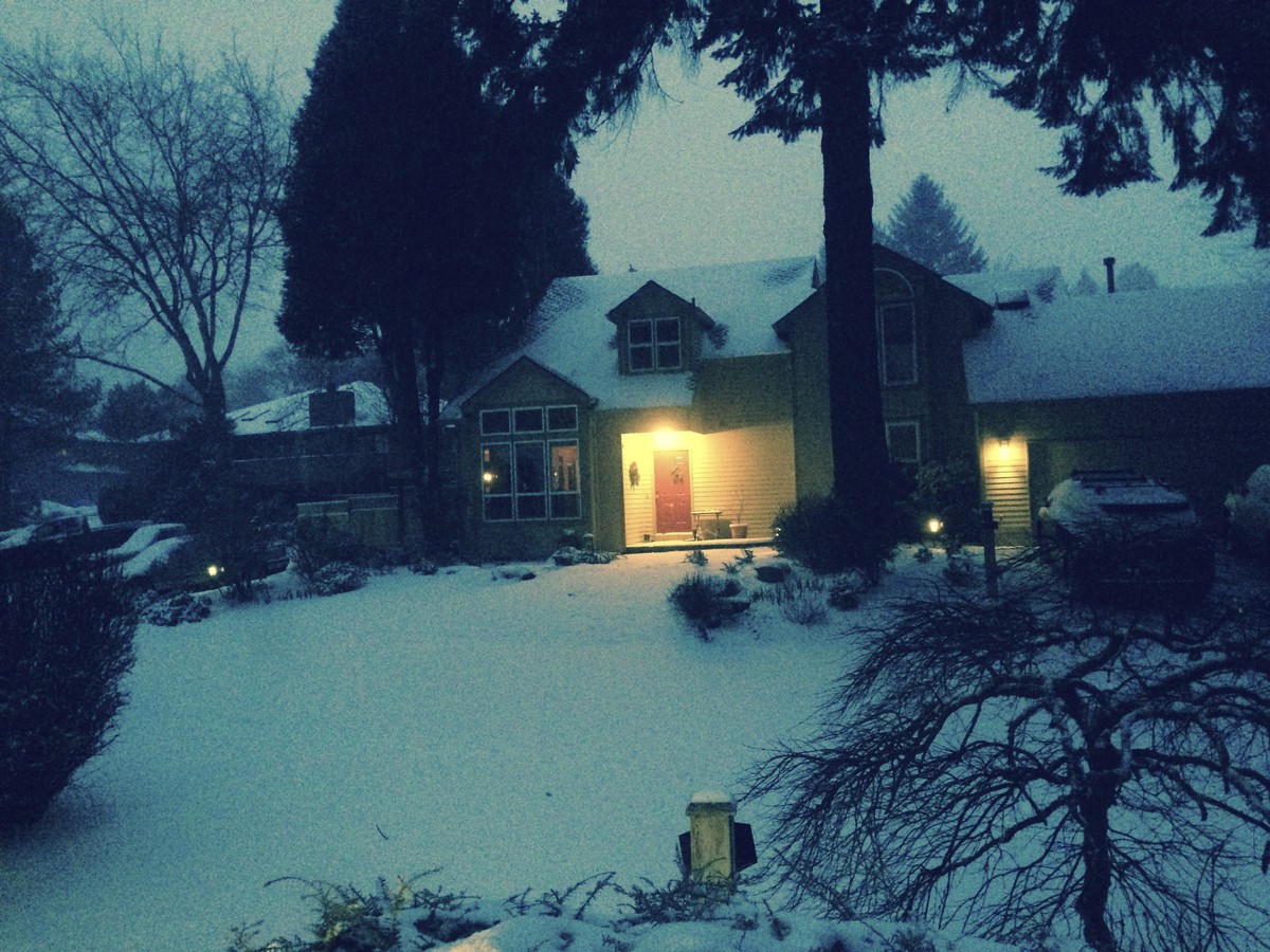 View onto a darkened snowy street; the house across the street has bright porch lights that shine warmly in the otherwise cold scene